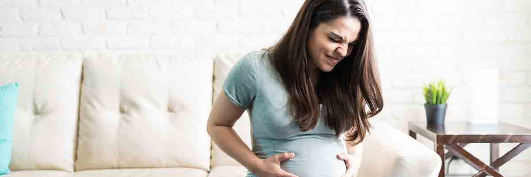 Attractive pregnant female grimacing with hands on baby bump while sitting at home